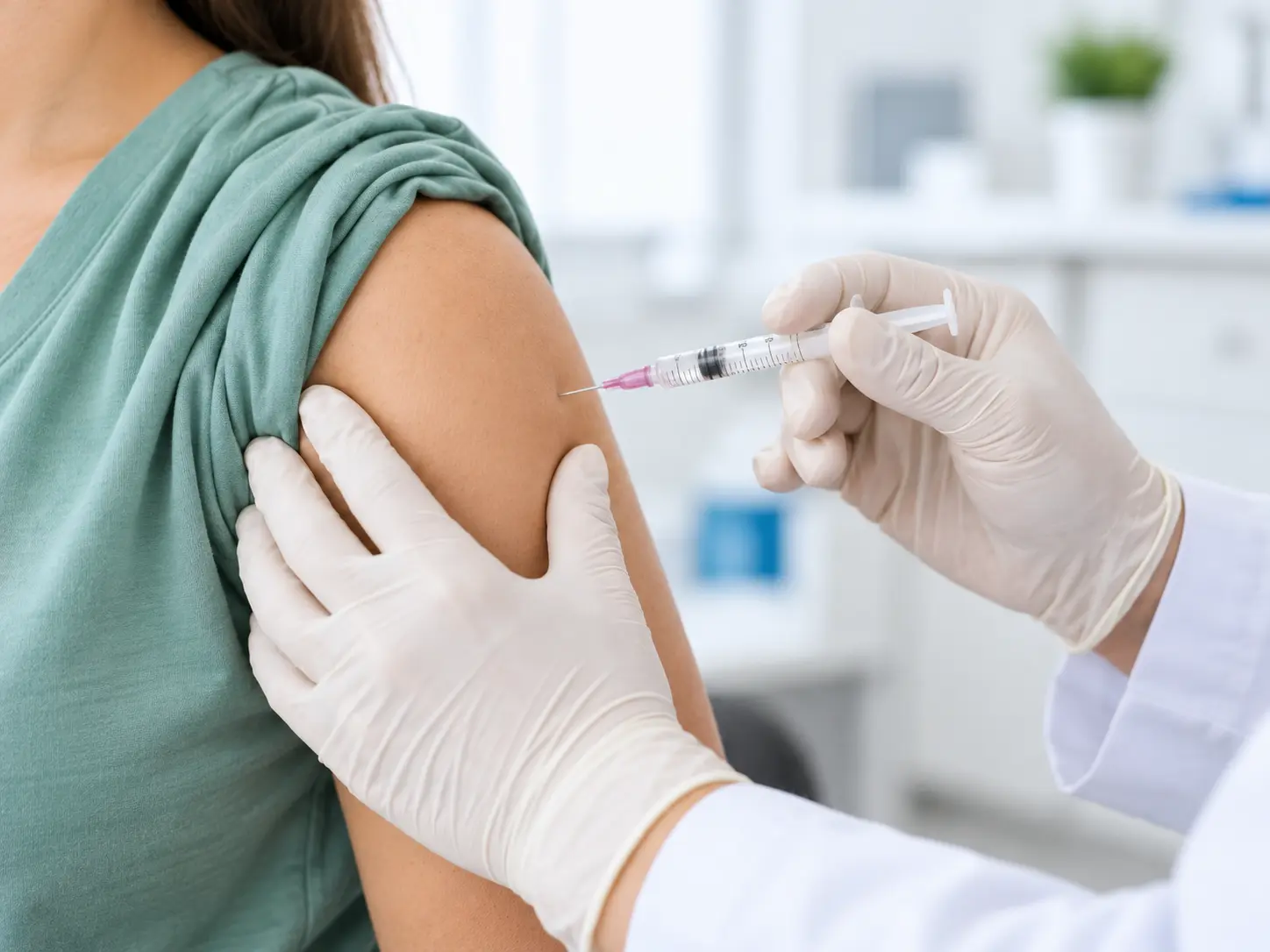 Healthcare professional wearing medical gloves positioning a syringe near a patient's upper arm in a clinical setting.
